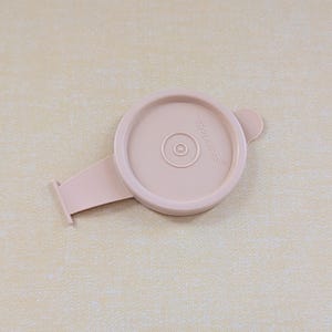May include: A light pink circular plastic lid featuring a small tab on the side and an attached flat rectangular handle extension. The word Tupperware is embossed on the top surface of the lid, which has a raised circular center design.