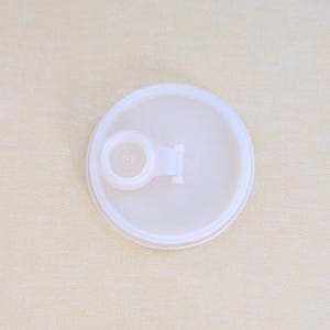 May include: A circular, translucent white plastic lid featuring a small, integrated spout with a hinged cap. The lid has a raised outer rim and a smooth, flat surface, designed to fit securely onto a container for liquid dispensing.