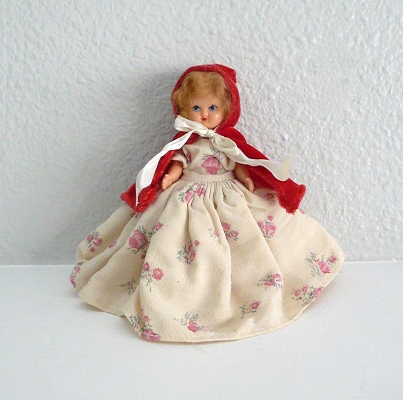 Puede incluir: Una mu&ntilde;eca vintage con una capa roja con capucha y un vestido blanco con estampado floral rosa.