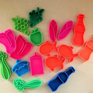 May include: A set of 14 colorful plastic cookie cutters shaped like various foods, including grapes, an apple, celery, a cupcake, a fish, a bottle of soda, and a steak.
