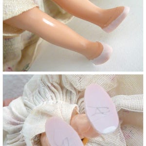 Puede incluir: Una mu&ntilde;eca vintage con un vestido blanco y zapatos rosas. Las piernas de la mu&ntilde;eca est&aacute;n dobladas en las rodillas y los zapatos son visibles. La mu&ntilde;eca lleva un vestido blanco con un estampado floral.