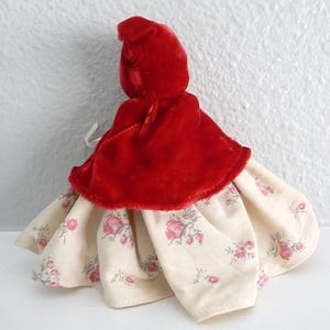 Puede incluir: Una capa de terciopelo rojo con capucha y un vestido blanco con estampado floral debajo. El vestido tiene una falda fruncida y la capa tiene una capucha puntiaguda.