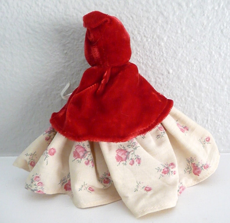 Puede incluir: Una capa de terciopelo rojo con capucha y un vestido blanco con estampado floral debajo. El vestido tiene una falda fruncida y la capa tiene una capucha puntiaguda.
