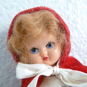 Puede incluir: Una mu&ntilde;eca vintage con cabello rubio que lleva una capa de terciopelo rojo con capucha y un lazo de cinta blanca. La mu&ntilde;eca tiene ojos azules y pesta&ntilde;as pintadas.