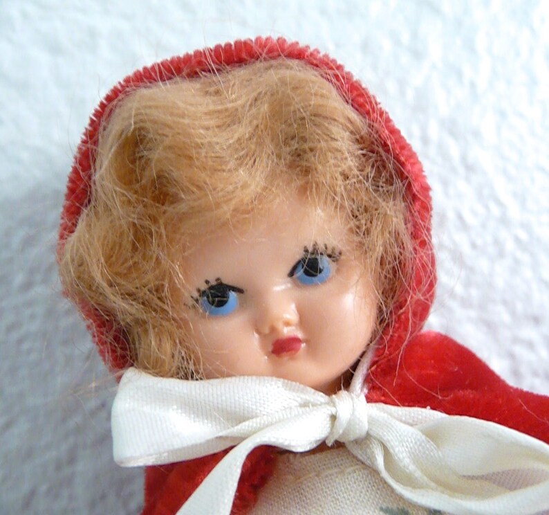 Puede incluir: Una mu&ntilde;eca vintage con cabello rubio que lleva una capa de terciopelo rojo con capucha y un lazo de cinta blanca. La mu&ntilde;eca tiene ojos azules y pesta&ntilde;as pintadas.