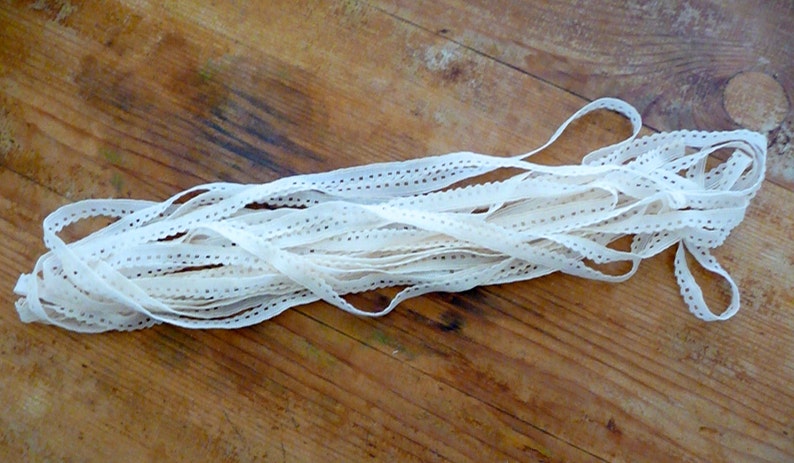 May include: A white cotton lace trim with a scalloped edge and small holes throughout. The lace is folded and piled on a wooden surface.