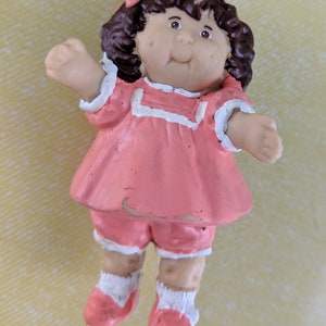 May include: A pink and white Cabbage Patch Kid doll figurine with brown hair and a pink bow. The doll is wearing a pink dress with white trim and pink shoes.