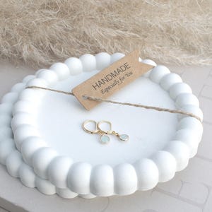 Könnte beinhalten: Zwei weiße, perlenbesetzte Dekotabletts mit einem Paar goldfarbener Ohrringe mit hellblauen Steinen. Ein kleines Etikett mit der Aufschrift "HANDMADE Especially for You" ist mit Schnur an den Tabletts befestigt. Der Hintergrund zeigt getrocknetes Ziergras.
