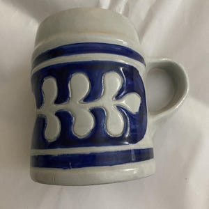 May include: A ceramic beer stein with a handle, featuring a light gray base and a dark blue floral design. The design is repeated around the body of the stein. The stein has a capacity of approximately 16 ounces.