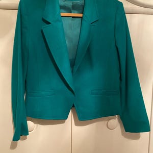 May include: Teal blazer with a notched lapel, two front pockets, and a single-button closure. The jacket is hanging on a wooden hanger. The interior lining is a lighter shade of teal. The blazer is a classic style.