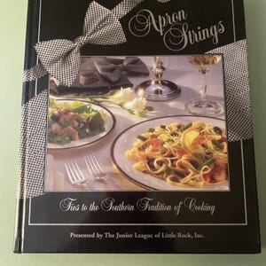 May include: A black hardcover cookbook titled Apron Strings: Ties to the Southern Tradition of Cooking, presented by The Junior League of Little Rock, Inc. The cover features a black and white gingham ribbon graphic and a photograph of pasta and salad.