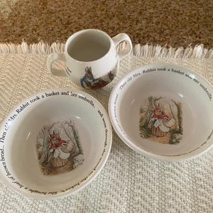 Peut inclure: Un mug en céramique blanche à deux anses et deux bols assortis illustrés d'un lapin en cape rouge portant un panier. Le bord des bols porte l'inscription : Then old Mrs. Rabbit took a basket and her umbrella.