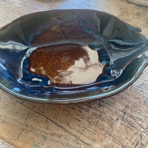 May include: An oval-shaped ceramic spoon rest featuring a glossy glaze with a deep brown center that transitions into dark blue and black tones along the curved edges. The surface has a smooth, reflective finish with visible color variations.