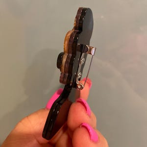May include: A black and brown brooch with a silver pin fastening. The brooch features a layered design with a glossy finish. The brooch is held in a hand with pink nail varnish.