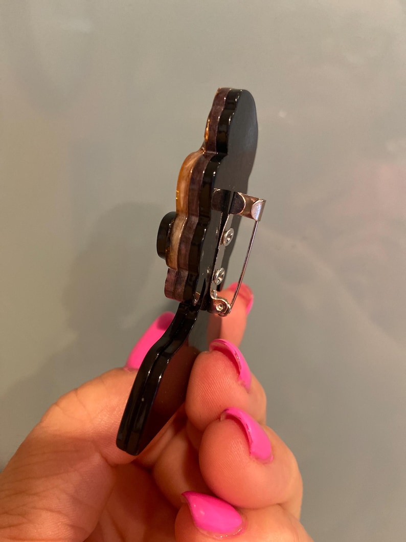 May include: A black and brown brooch with a silver pin fastening. The brooch features a layered design with a glossy finish. The brooch is held in a hand with pink nail varnish.