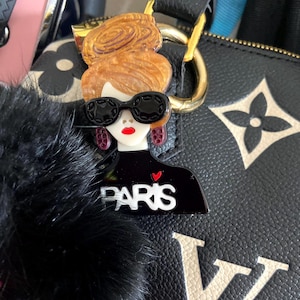 May include: A black and white acrylic charm depicting a woman with sunglasses, red lips, and a bun. The charm is attached to a black bag with a gold clasp and a black fur pom-pom. The word "PARIS" is printed on the charm.