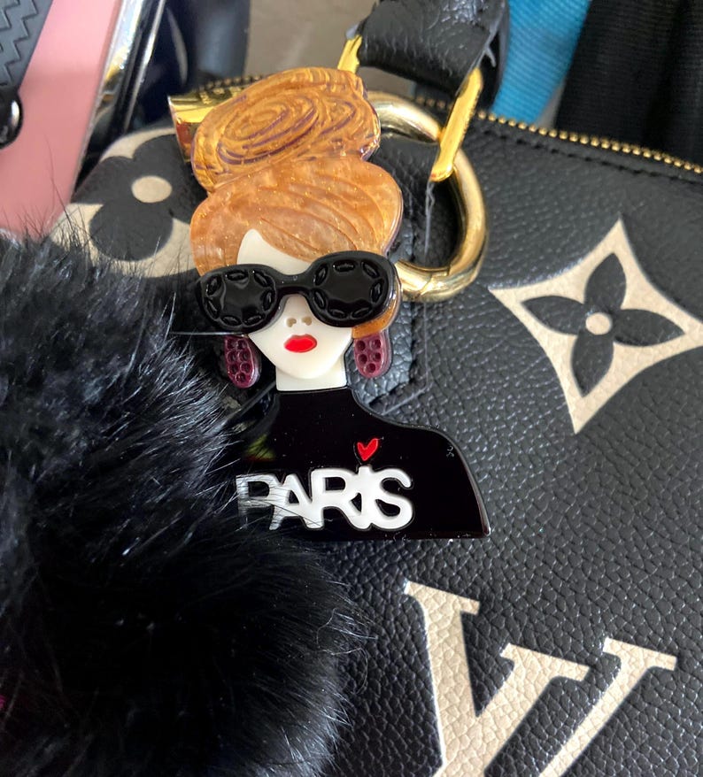 May include: A black and white acrylic charm depicting a woman with sunglasses, red lips, and a bun. The charm is attached to a black bag with a gold clasp and a black fur pom-pom. The word "PARIS" is printed on the charm.
