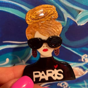 May include: A brooch depicting a woman with a golden updo, black sunglasses, and red lipstick. The black top has the word "PARIS" in white with a red heart. The brooch is set against a blue and white backdrop.