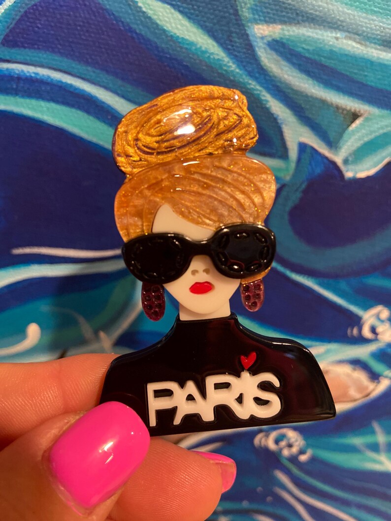 May include: A brooch depicting a woman with a golden updo, black sunglasses, and red lipstick. The black top has the word "PARIS" in white with a red heart. The brooch is set against a blue and white backdrop.