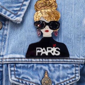 May include: A fashionable brooch depicting a woman with sunglasses and a gold bun. The black top displays the word "PARIS" in white. The brooch is pinned to a blue denim jacket.