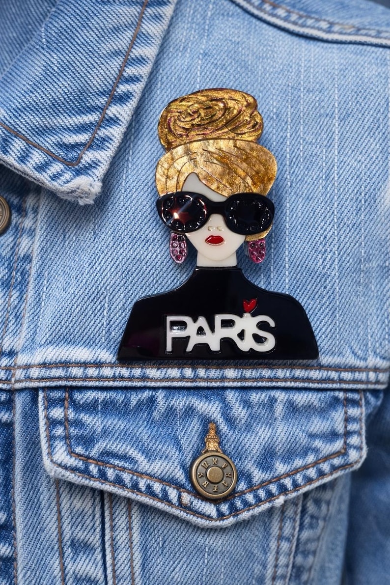 May include: A fashionable brooch depicting a woman with sunglasses and a gold bun. The black top displays the word "PARIS" in white. The brooch is pinned to a blue denim jacket.