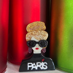 May include: A black and white brooch featuring a stylized woman with sunglasses and the word "PARIS". The brooch has a gold-coloured hairstyle and red lips. The woman's top has a small red heart. The background includes red and green cylindrical objects.