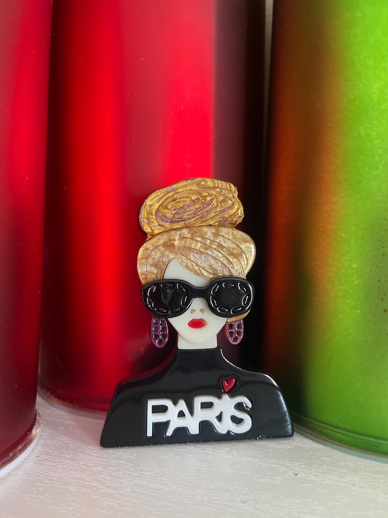 May include: A black and white brooch featuring a stylized woman with sunglasses and the word "PARIS". The brooch has a gold-coloured hairstyle and red lips. The woman's top has a small red heart. The background includes red and green cylindrical objects.