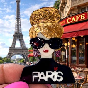 May include: A black and white brooch depicting a woman with sunglasses and a bun, with the word "PARIS". The Eiffel Tower and a cafe are visible in the background. The brooch is a fashion accessory.