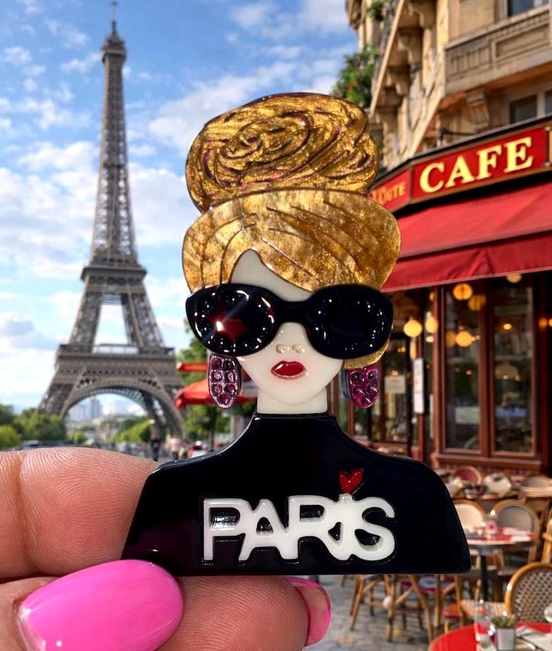 May include: A black and white brooch depicting a woman with sunglasses and a bun, with the word "PARIS". The Eiffel Tower and a cafe are visible in the background. The brooch is a fashion accessory.