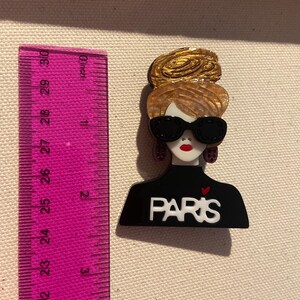 May include: A fashion-themed brooch featuring a stylized woman with sunglasses and a gold updo. The black top has the word "PARIS" in white. The brooch is approximately 5 cm in height.