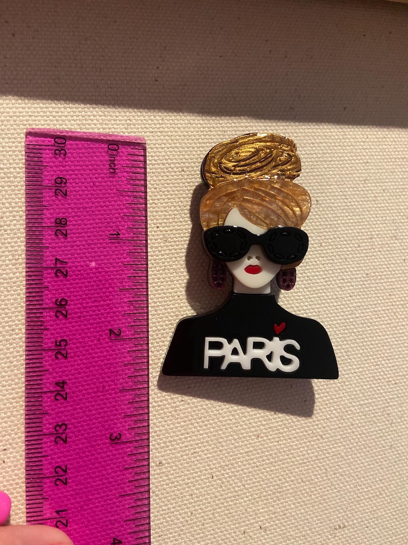 May include: A fashion-themed brooch featuring a stylized woman with sunglasses and a gold updo. The black top has the word "PARIS" in white. The brooch is approximately 5 cm in height.
