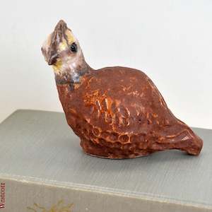 May include: A ceramic bird figurine with a brown body and a head with yellow, white, and brown details. The bird is perched on a book. The figurine has a textured surface.