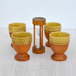 May include: A set of four egg cups with wooden bases and yellow ceramic tops featuring a textured pattern, arranged around a central wooden hourglass with white sand. The egg cups have a rounded bowl shape and a pedestal base, while the hourglass.