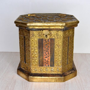 May include: An antique, octagonal chest with ornate gold-colored metalwork and a hinged lid. The chest features intricate floral and geometric patterns, with a central panel of wood and copper accents. A brass clasp secures the lid.