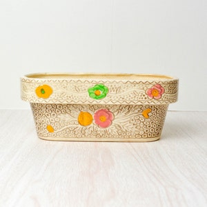 May include: A white ceramic planter with a brown floral design. The planter has a rectangular shape and features a raised rim with a scalloped edge. The floral design includes pink, green, and orange flowers.