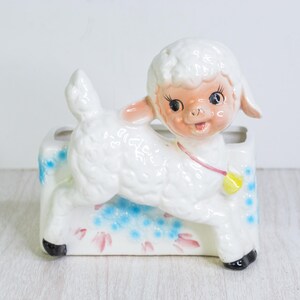 May include: A vintage ceramic lamb planter with a white body, pink ears, and a smiling face. The planter has a rectangular base decorated with blue and pink floral accents. Black hooves and a yellow bell complete the design.