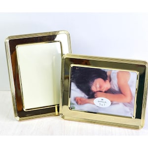 May include: Two rectangular picture frames with a polished gold-tone finish and a stepped border design. One frame is oriented vertically and the other horizontally, containing a photograph of a sleeping child with a Hallmark 5 x 7 label.