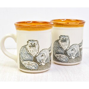 May include: Two off-white ceramic mugs featuring a brown glazed rim and a detailed illustration of long-haired cats. One mug shows a cat sitting next to a kitten playing with a ball, while the other shows a cat and a kitten interacting together.
