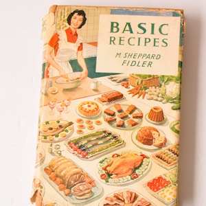 May include: A vintage cookbook titled "Basic Recipes" by M. Sheppard Fidler. The cover features a woman in a white apron preparing food in a kitchen, surrounded by a colorful spread of various dishes, including roasted chicken, fish, and desserts.