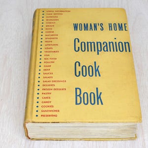 May include: A vintage cookbook titled "Woman's Home Companion Cook Book" with a yellow cover and blue text. The book's spine lists various food categories. The book is on a light wood surface.