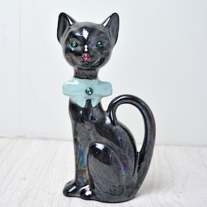 May include: A black ceramic cat figurine with iridescent finish. The cat has green eyes, a red nose, and a light blue bow tie with a gem. The cat is sitting upright with its tail curled.