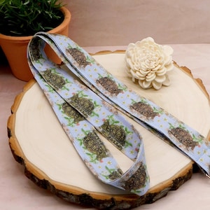 May include: A light blue lanyard with a repeating pattern of turtles and flowers. The lanyard is made of a soft fabric and has a metal clasp at the end. The lanyard is laying on a wooden surface.