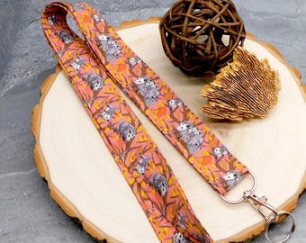 May include: A pink lanyard with a repeating pattern of opossums in trees with orange and yellow leaves. The lanyard has a silver-colored clasp and key ring. The lanyard is lying on a wooden surface.