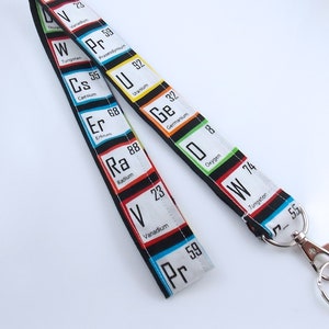 May include: A black and white lanyard with colorful squares featuring the chemical symbols and names of elements, such as Vanadium, Tungsten, Caesium, Erbium, Radium, Uranium, Germanium, Oxygen, and  Tungsten. The lanyard has a silver metal clasp.
