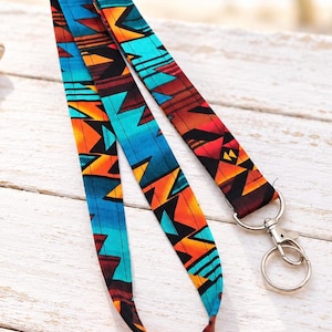 May include: A fabric lanyard featuring a repeating geometric pattern in vibrant shades of turquoise, orange, red, and black. The strap is attached to a silver-toned metal swivel lobster clasp and a circular split ring for holding keys or ID badges.