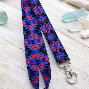 Op de afbeelding: Een stoffen keycord met een herhalend geometrisch patroon in blauwe, rode en zwarte tinten. De band is in een lus gevouwen en bevestigd aan een zilverkleurige metalen draaibare karabijnhaak en een ronde sleutelring voor sleutels of pasjes.