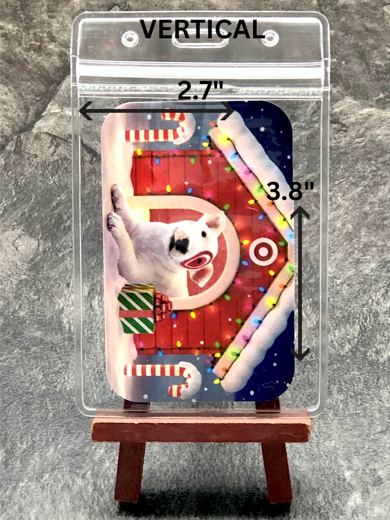 May include: A clear plastic badge holder with a white dog wearing a red Santa hat and a red and white candy cane in a red and white Christmas house with lights. The holder is 2.7 inches wide and 3.8 inches tall.