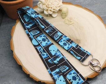 May include: A black lanyard with a repeating pattern of light blue x-ray images of skulls and bones. The lanyard has a silver-colored metal clasp. The lanyard is displayed on a wooden slice.