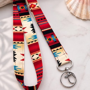 Op de afbeelding: Een stoffen lanyard met een herhalend geometrisch patroon in tinten rood, crème, zwart en felblauw. De band is bevestigd aan een zilverkleurige metalen draaibare karabijnhaak en een ronde sleutelring voor het bevestigen van sleutels of pasjes.