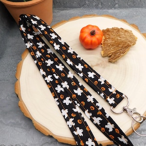 May include: A black fabric lanyard featuring a repeating pattern of white ghosts and small orange pumpkins. The strap is stitched with orange thread and includes a silver-toned metal swivel lobster clasp and a circular split ring at the end.